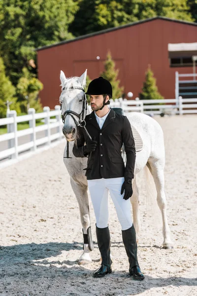 Schöner männlicher Reiter, der neben Pferd steht und beim Pferdeclub wegschaut — Stockfoto