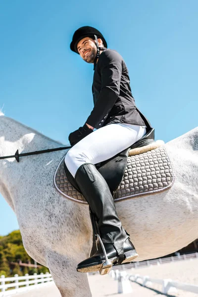 Bajo ángulo vista de sonriente guapo macho ecuestre sentado a caballo en el club de caballos - foto de stock