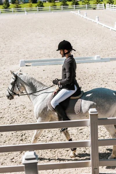 Vista laterale di bel maschio equestre in casco da equitazione, giacca nera e pantaloni bianchi a cavallo al club di cavalli — Foto stock