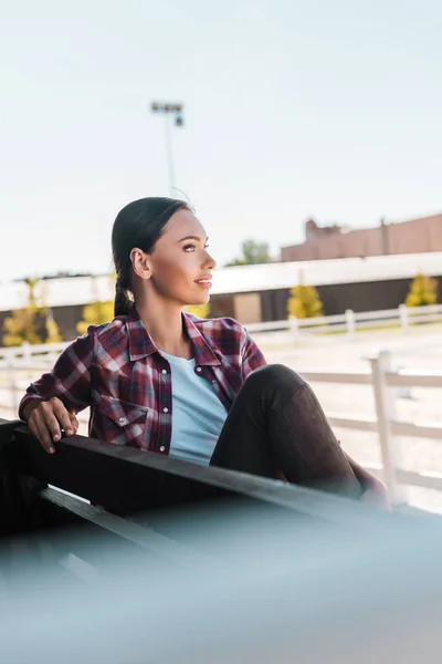 Vue latérale d'une jolie cow-girl en chemise à carreaux assise sur un banc au stade du ranch et regardant ailleurs — Photo de stock