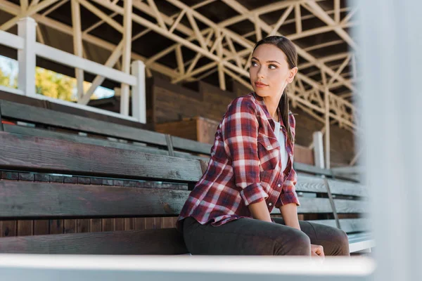 Beautiful cowgirl in checkered shirt sitting on bench at ranch stadium and looking away — Stock Photo