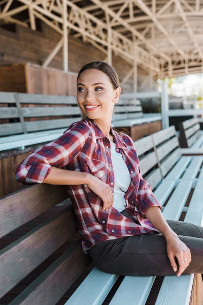 Femme attrayante souriante en chemise à carreaux assis sur le banc au stade du ranch et regardant loin — Photo de stock