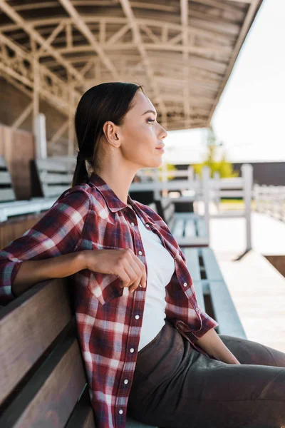Вид сбоку красивой девушки в черной рубашке, сидящей на скамейке на стадионе ранчо и отводящей взгляд — стоковое фото