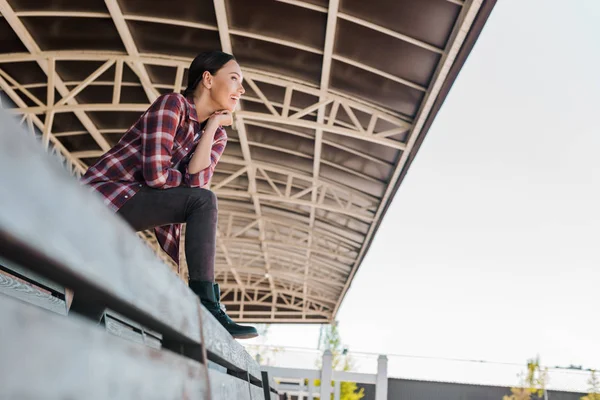 Nivel de superficie de la mujer atractiva en camisa a cuadros sentado en el banco en el estadio rancho y mirando hacia otro lado - foto de stock