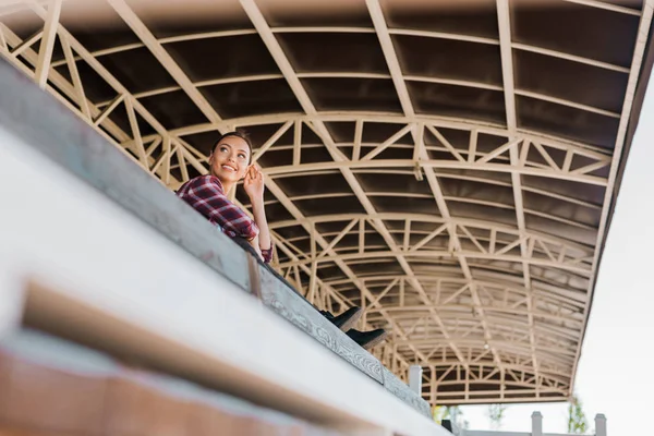 Low angle view of attractive cowgirl in checkered shirt sitting on bench at ranch stadium — Stock Photo