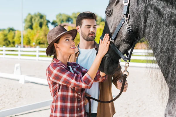 Cowboy and cowgirl in casual clothes palming black horse at ranch — Stock Photo
