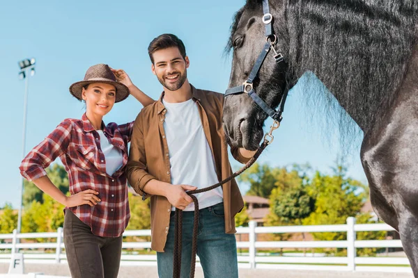 Hembras sonrientes y equestrians masculinos de pie cerca del caballo en el rancho y mirando a la cámara - foto de stock