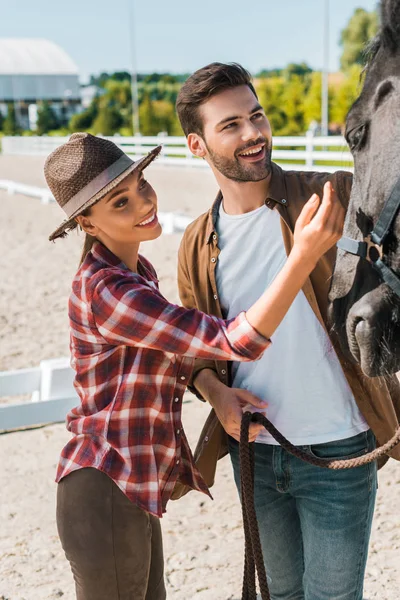 Sorridente cowboy e cowgirl palming cavallo nero al ranch — Foto stock