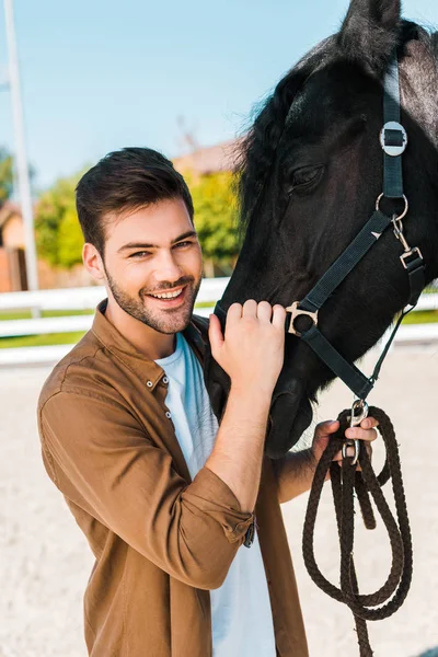 Smiling handsome male equestrian holding horse halter and looking at camera at ranch — Stock Photo