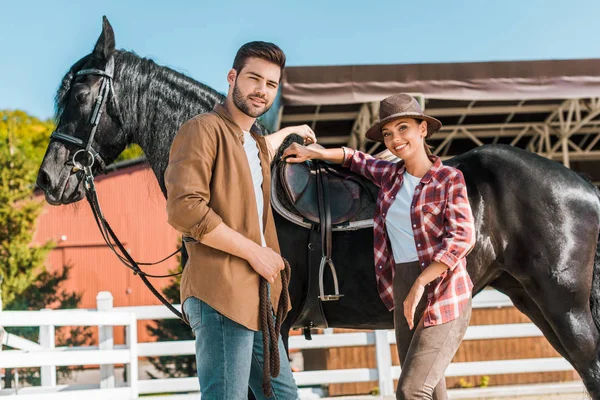 Low angle view of cowboy and cowgirl standing near horse at ranch and looking at camera — Stock Photo