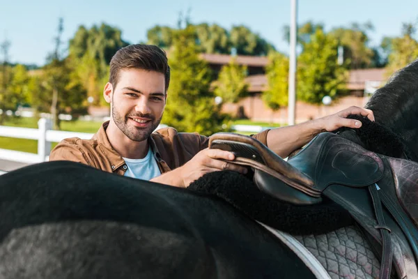 Lächelnd schöner männlicher Pferdesattel auf Ranch repariert — Stockfoto