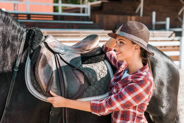 Vista lateral de sorrir atraente fêmea equestre fixação cavalo sela no rancho — Fotografia de Stock