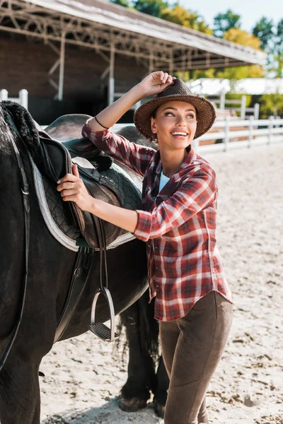 Atraente sorrindo equestre fixação cavalo sela e tocando chapéu na fazenda — Fotografia de Stock