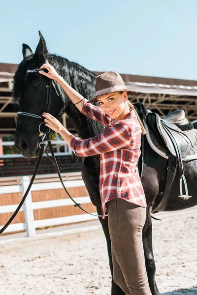 Attractive cowgirl fixing horse halter at ranch and looking at camera — Stock Photo