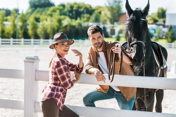 Vaquero sonriente y vaquera de pie cerca de la cerca con el caballo en el rancho - foto de stock