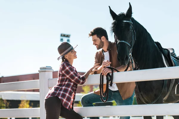 Sorridentes equestres femininos e masculinos em pé perto de cerca com cavalo e olhando um para o outro na fazenda — Fotografia de Stock