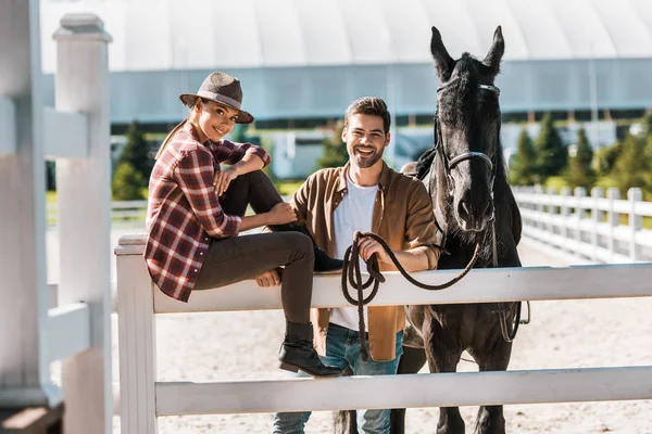 Cavallerizza sorridente seduta sulla recinzione, collega in piedi vicino alla recinzione con cavallo al ranch — Foto stock