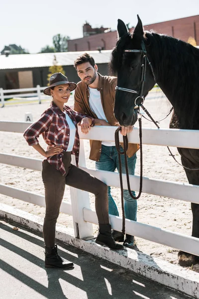 Female and male equestrians standing near fence with horse at ranch and looking at camera — Stock Photo