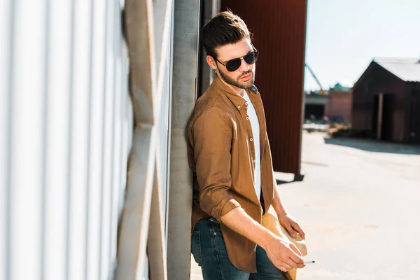 Handsome cowboy in sunglasses holding cigarette and leaning on wall at ranch — Stock Photo