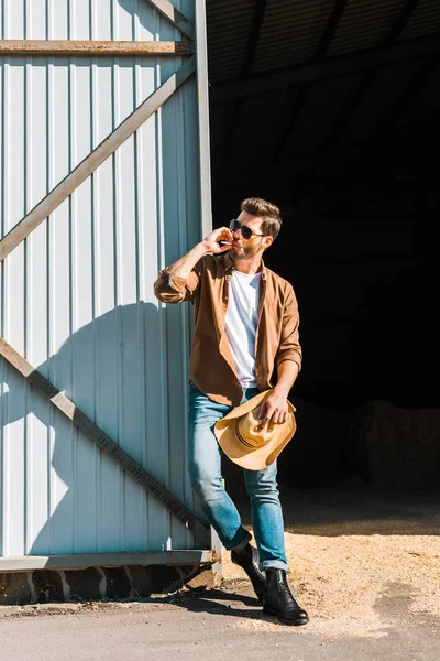 Hombre guapo en gafas de sol fumar cigarrillo, sosteniendo sombrero y apoyándose en la pared en el rancho - foto de stock