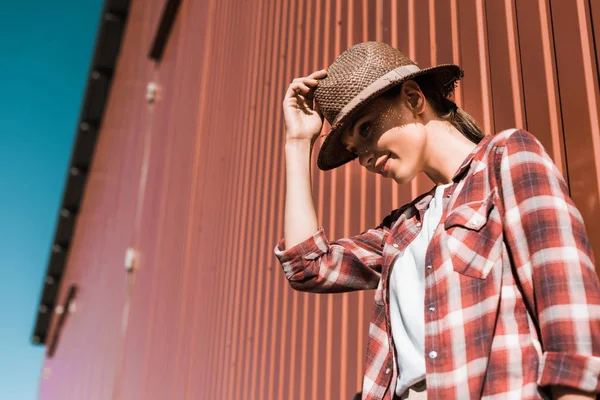Низкий угол зрения привлекательной женщины в клетчатой рубашке и шляпе опираясь на коричневую стену на ранчо — Stock Photo