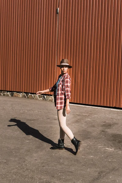 Attractive cowgirl in checkered shirt and hat walking near brown building at ranch — Stock Photo