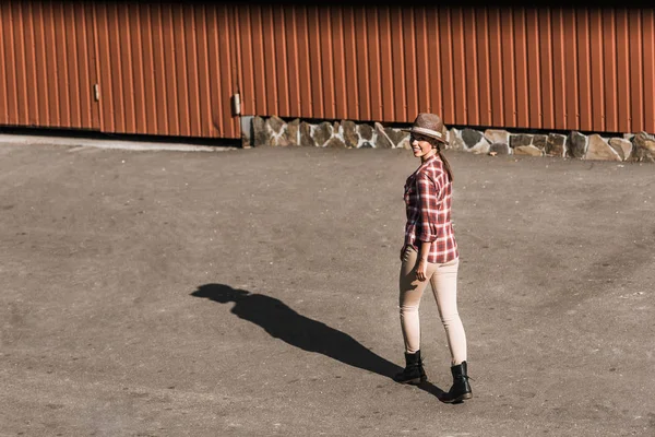 Jolie cowgirl souriante en chemise à carreaux et chapeau marchant près du bâtiment brun au ranch — Photo de stock