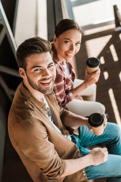 Visão de alto ângulo de homem sorridente e mulher sentada com café para ir ao rancho e olhar para a câmera — Fotografia de Stock