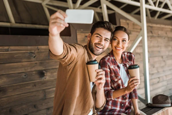 Hombre y mujer sonrientes tomando selfie con teléfono inteligente y sosteniendo tazas de café desechables en el rancho - foto de stock