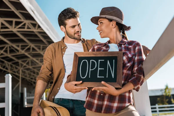 Stylish cowboy and cowgirl in casual clothes holding open sign at ranch and looking at each other — Stock Photo