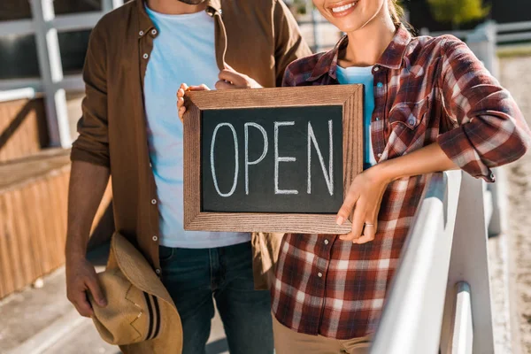 Cropped image of stylish equestrians in casual clothes holding open sign at ranch — Stock Photo