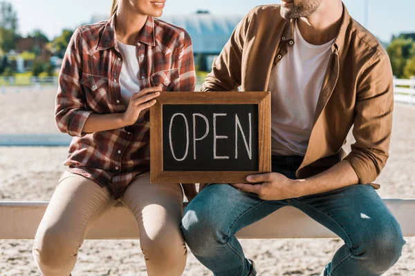 Cropped image of stylish equestrians in casual clothes sitting on fence and holding open sign at ranch — Stock Photo