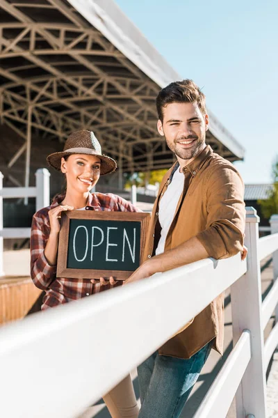 Stylish cowboy and cowgirl in casual clothes standing near fence and holding open sign at ranch — Stock Photo