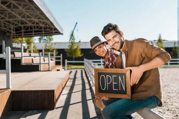 Smiling stylish equestrians in casual clothes sitting on fence, holding open sign and looking at camera at ranch — Stock Photo