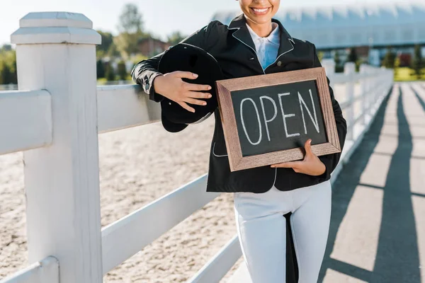 Cropped image of smiling equestrian leaning on fence and holding open sign at horse club — Stock Photo