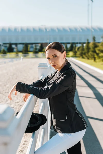 Attrayant équestre féminin appuyé sur la clôture et regardant la caméra au club de chevaux — Photo de stock