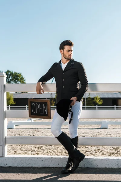 Bonito masculino equestre inclinação no cerca e segurando sinal aberto no cavalo clube — Fotografia de Stock