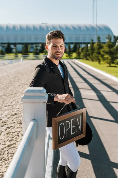 Schöner lächelnder Reiter lehnt am Zaun, hält offenes Schild und blickt in die Kamera auf der Ranch — Stockfoto