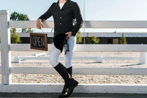 Cropped image of male equestrian leaning on fence and holding open sign at horse club — Stock Photo