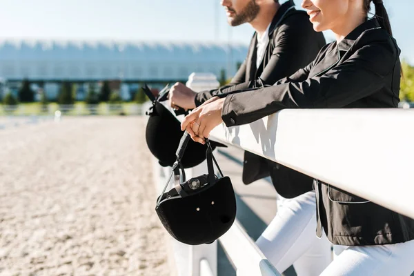 Cropped image of smiling equestrians in professional apparel leaning on fence and holding riding helmets at ranch — Stock Photo