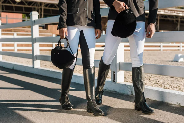 Imagen recortada de equestrians en ropa profesional caminando cerca de valla y sosteniendo cascos de montar en el rancho — Stock Photo