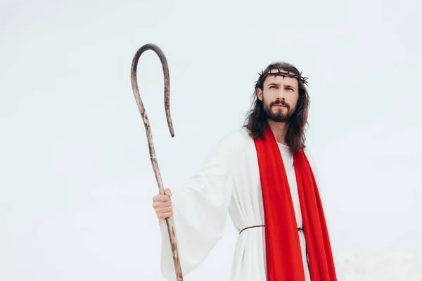 Low angle view of Jesus in robe, red sash and crown of thorns standing with wooden staff in desert — Stock Photo