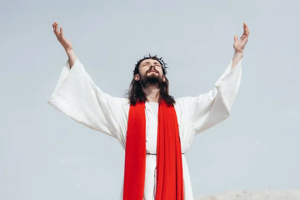 Jesus de roupão, faixa vermelha e coroa de espinhos de pé com as mãos levantadas e orando no deserto — Fotografia de Stock