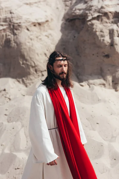 Handsome Jesus in robe, red sash and crown of thorns walking with closed eyes in desert — Stock Photo