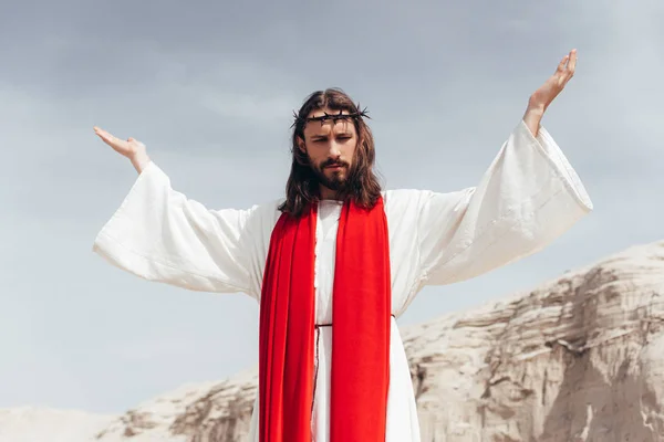 Low angle view of Jesus in robe, red sash and crown of thorns standing with raised hands in desert — Stock Photo