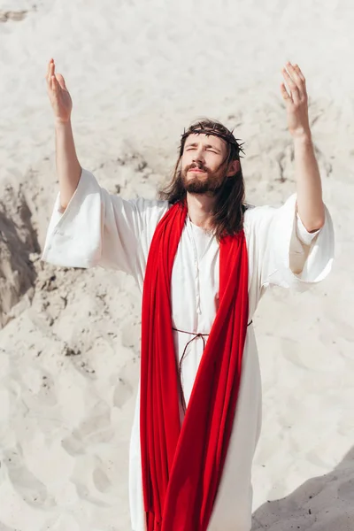 Иисус в мантии, красной ленте и терновом венце, стоя с поднятыми руками и молясь в пустыне — стоковое фото