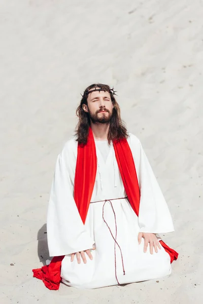 Jesus in robe, red sash and crown of thorns standing on knees with closed eyes and praying in desert — Stock Photo