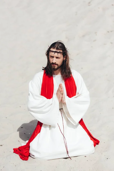 Jesús con túnica, faja roja y corona de espinas de rodillas y orando en el desierto, mirando hacia otro lado — Stock Photo