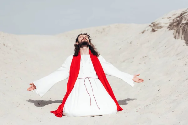 Jesus in robe, red sash and crown of thorns standing on knees with open arms in desert — Stock Photo