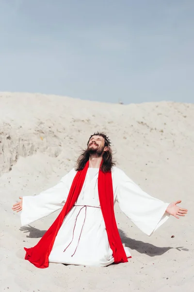 Happy Jesus in robe, red sash and crown of thorns standing on knees with open arms in desert — Stock Photo